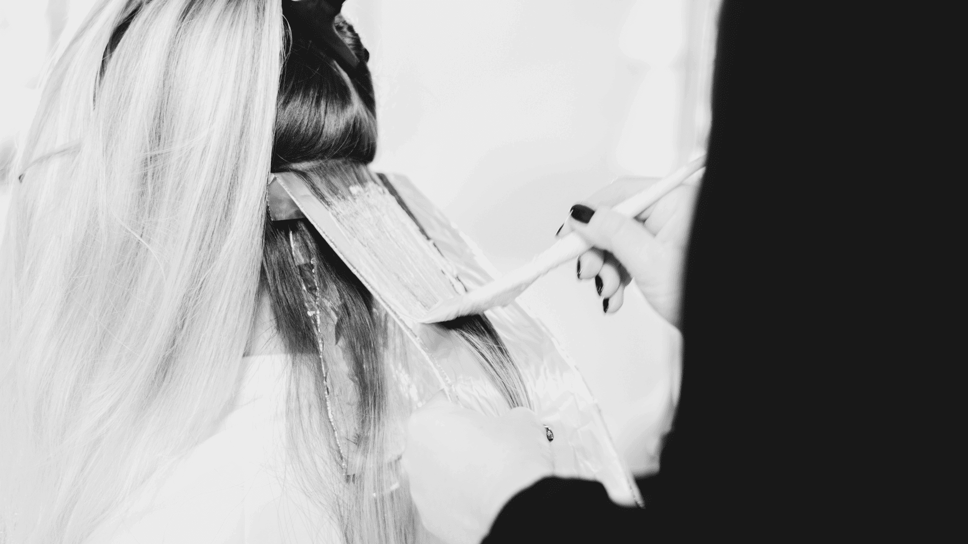 Hairdresser applying hair dye to client's blonde hair with a brush in a salon.