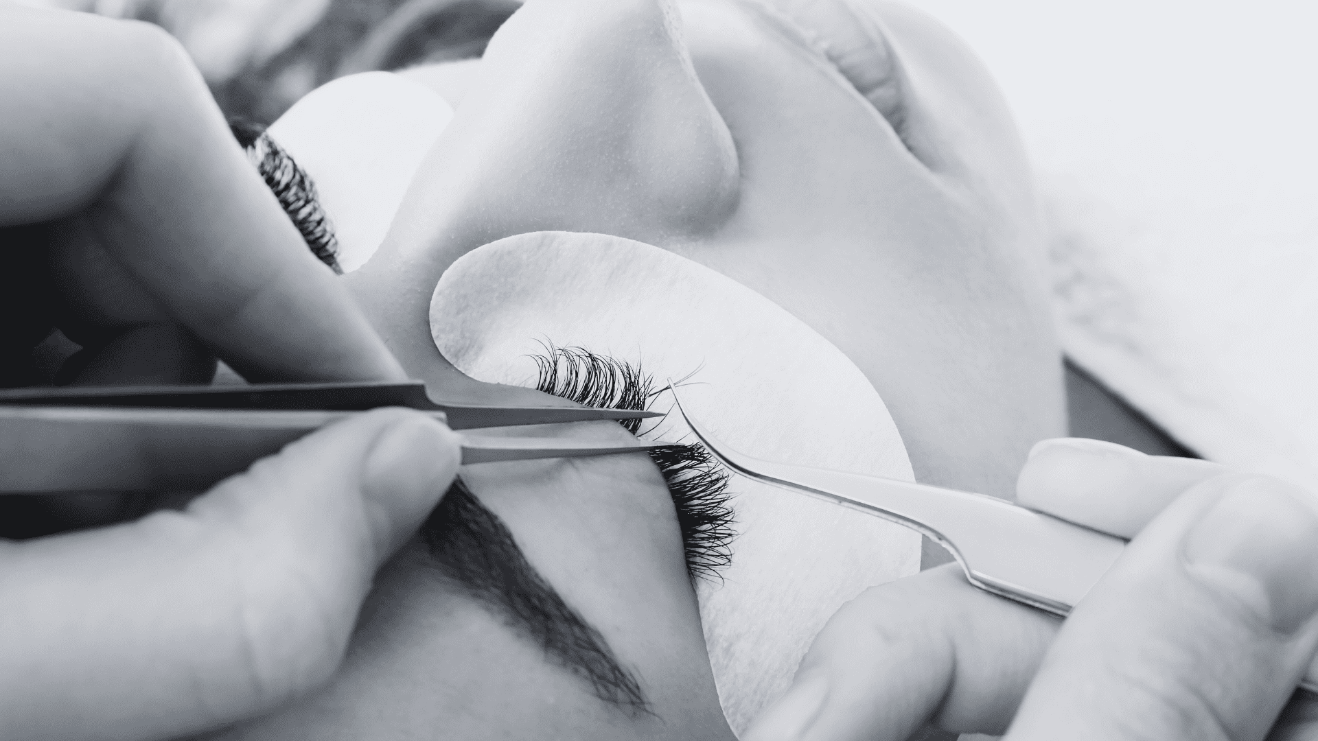 Close-up of a person getting eyelash extensions applied with tweezers and eye pads.