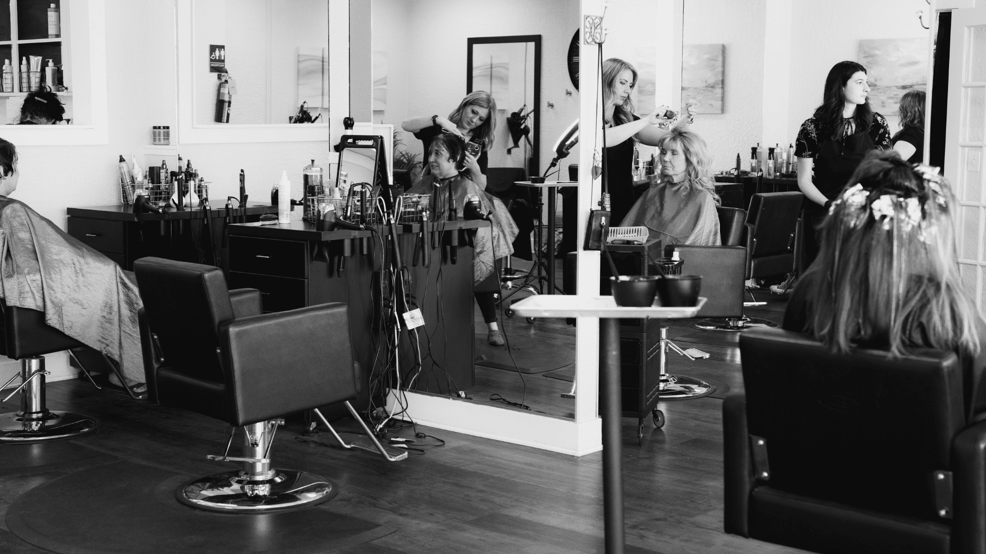 Stylists working on clients' hair in a busy salon, with mirrors and various hair products visible.